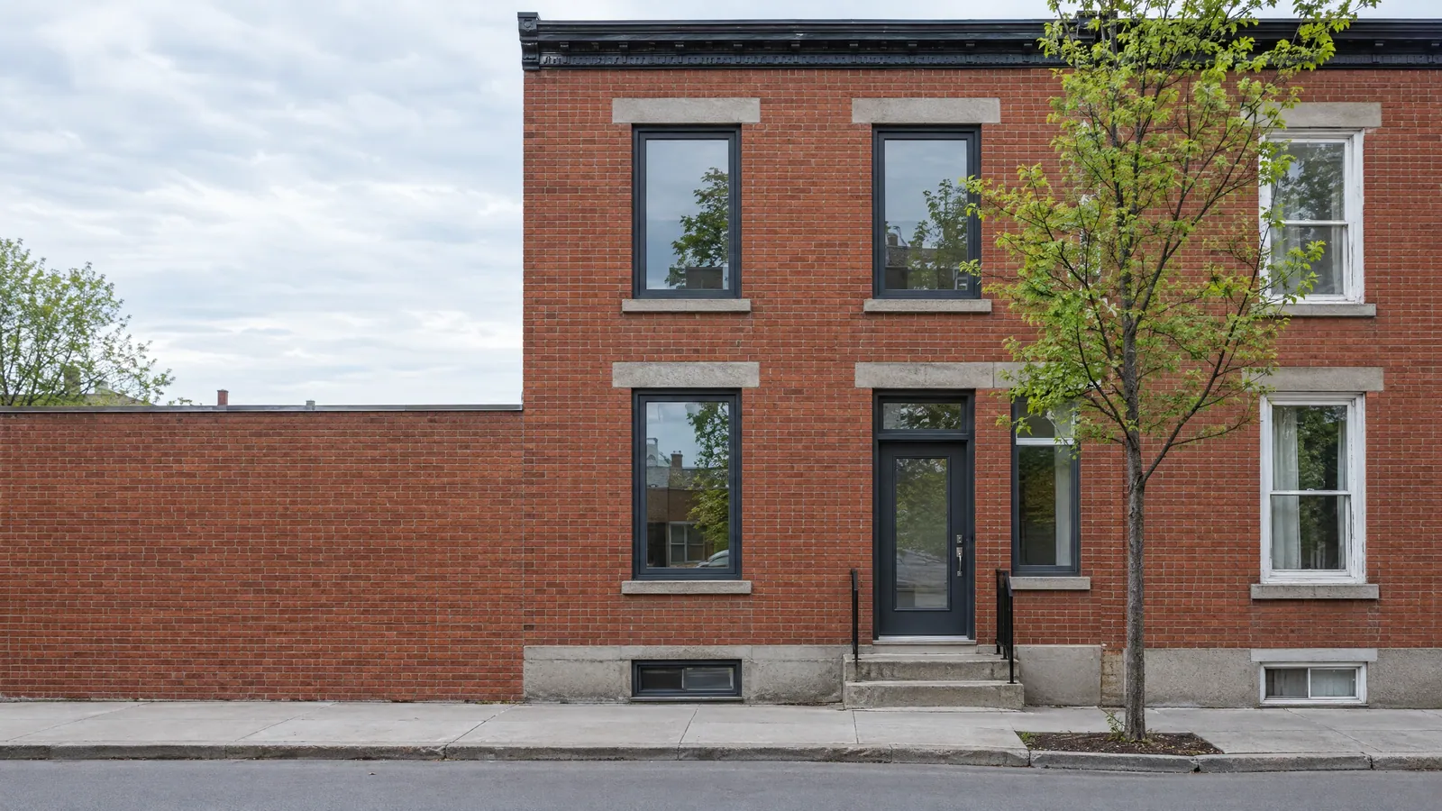 Red-brick row house with new anthracite hybrid windows and a matching door with transom, beside a neighbour's older white hung windows