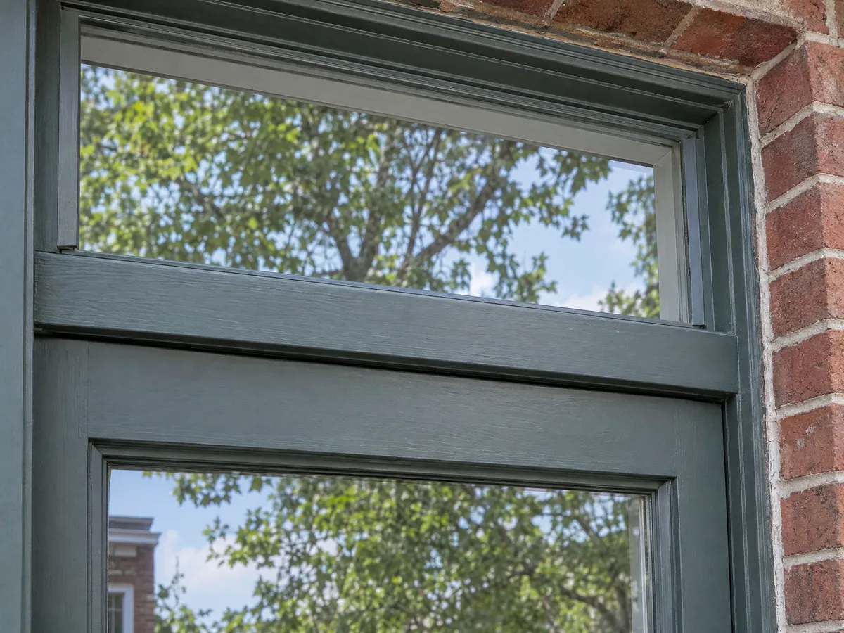 Close-up of a grey-green painted transom above a wood balcony door, showing the head jamb between the two panes of glass against red brick
