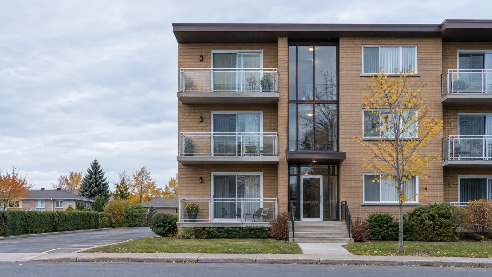 Three-storey tan-brick walk-up with glass-railed balconies and a sliding patio door on each floor, autumn trees along the street