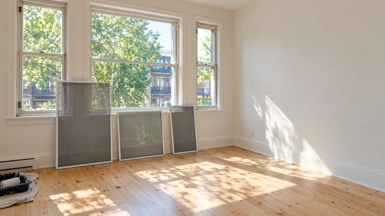 Three new window screens leaning against the wall beneath tall hung windows in an empty, freshly painted flat with wood floors