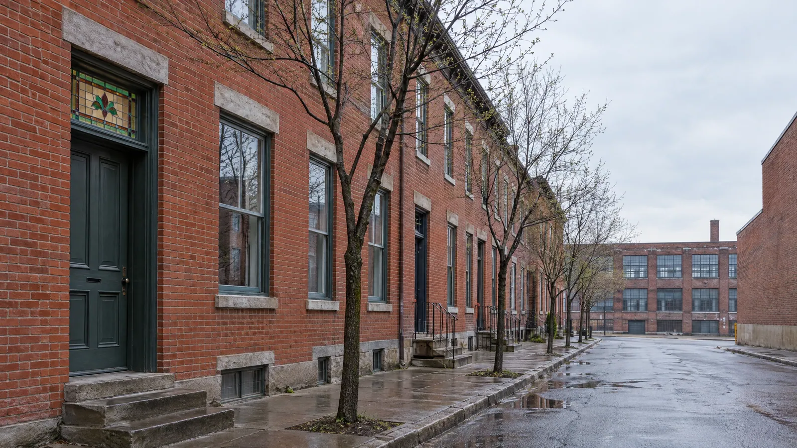 Wet street of red-brick row houses with tall wood windows and a dark green door under a stained-glass transom, bare young trees, and an old brick factory building at the end of the street