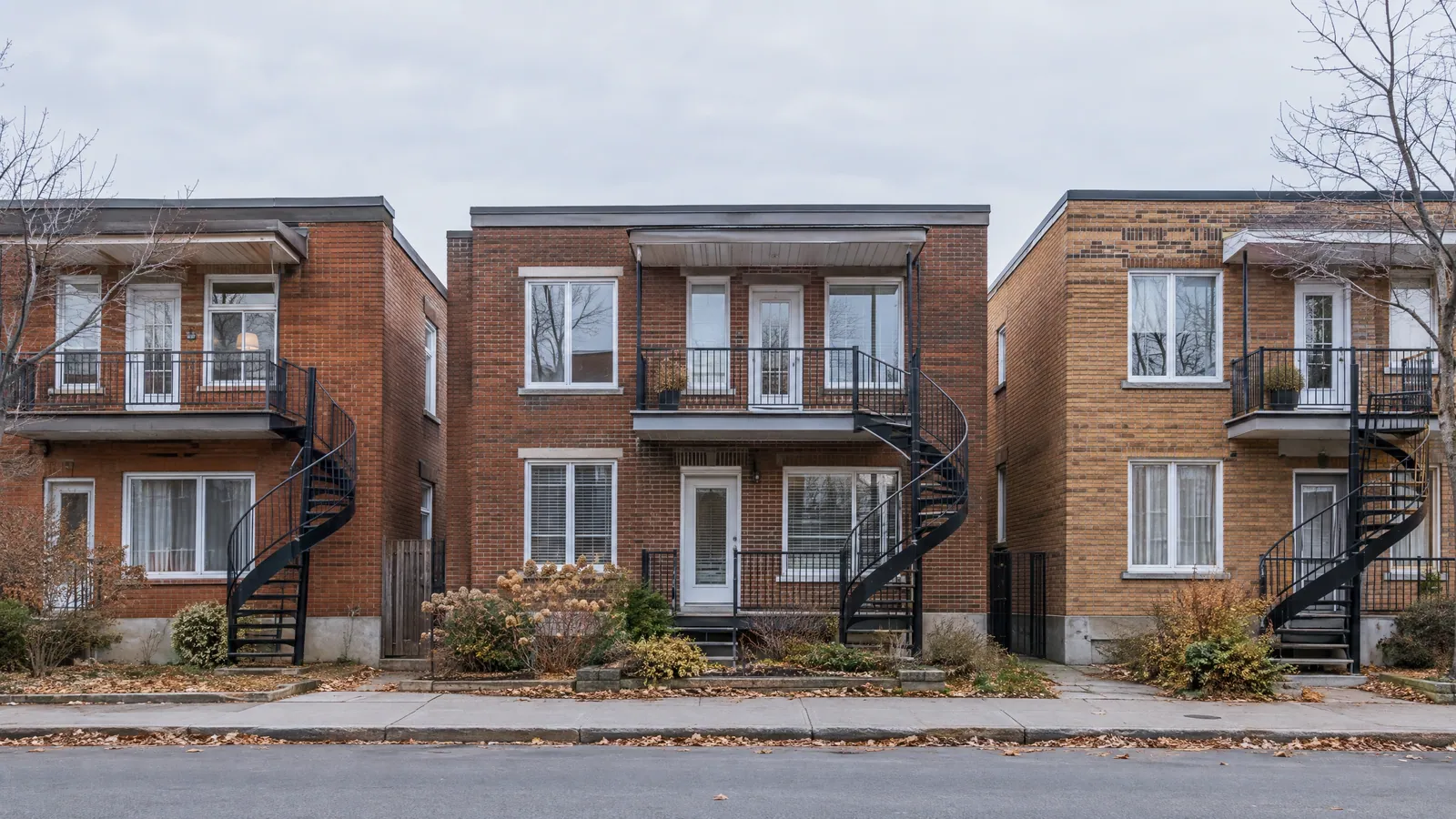 Three brick duplexes on a grey November day, each with a black spiral staircase rising to a second-floor balcony and door, bare trees and fallen leaves