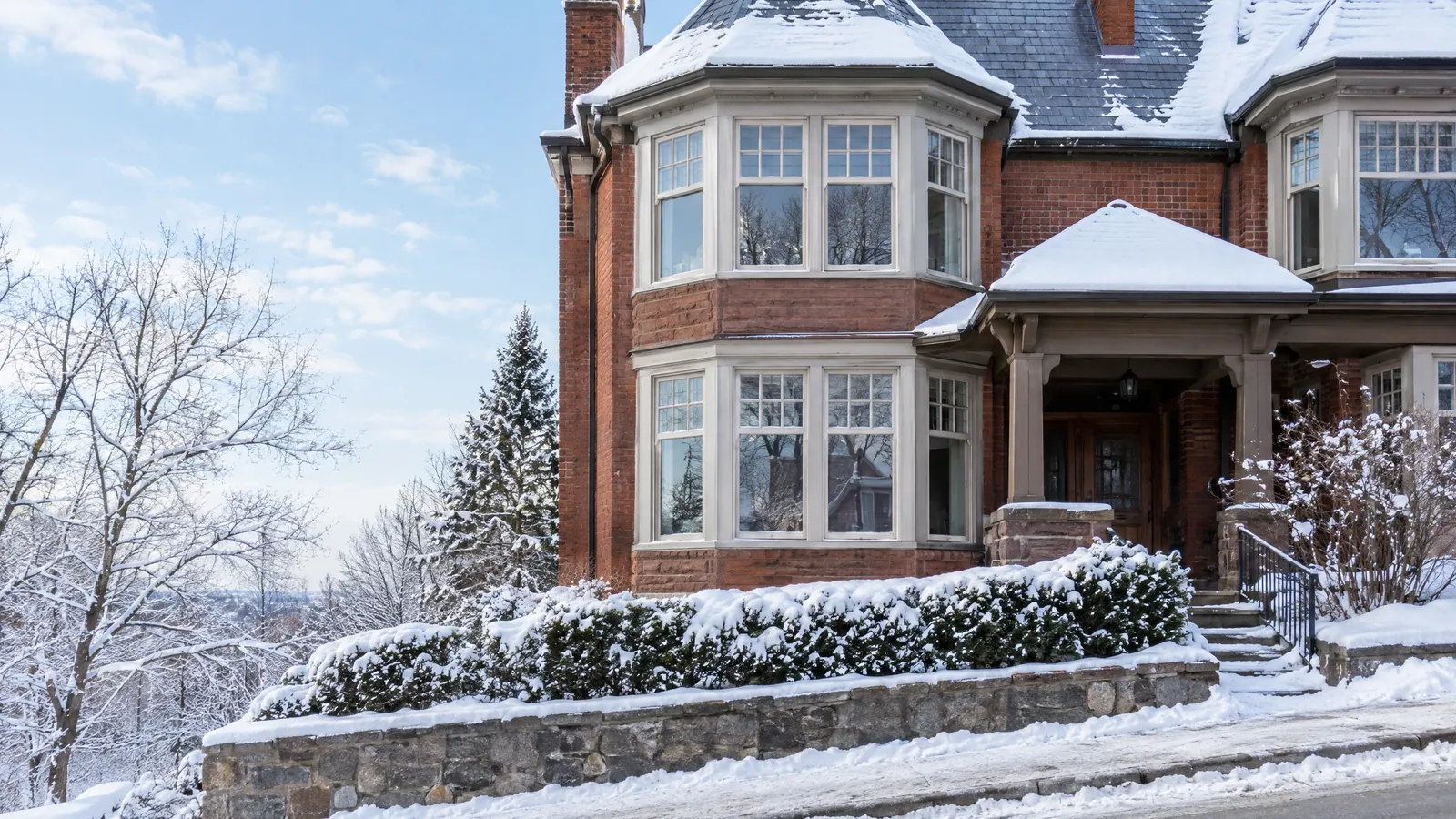 Red-brick semi-detached house with two-storey bay windows of divided wood sash, a covered porch and a stone garden wall under fresh snow